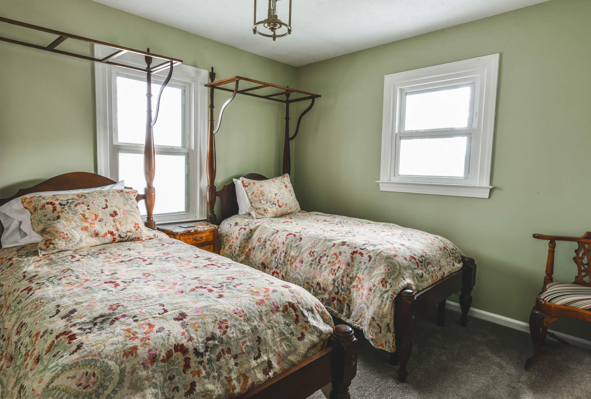 A small bedroom with two wooden twin beds, floral bedding, a wooden nightstand in between, two windows, green walls, and a striped chair in the corner.