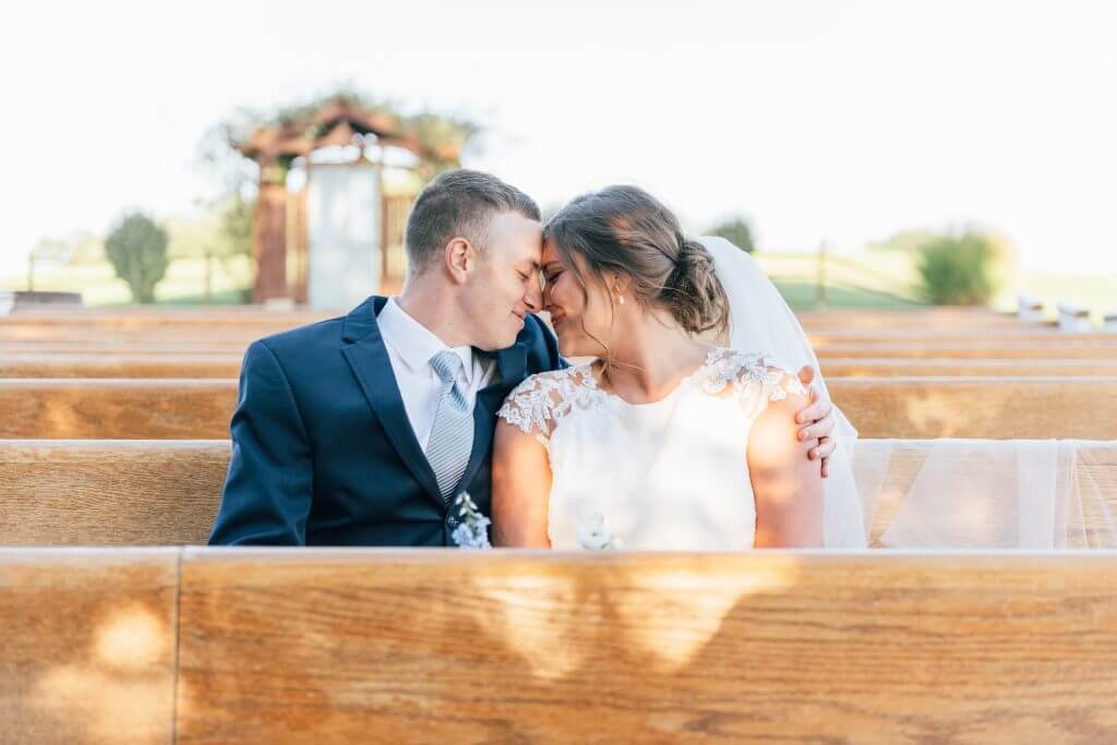 experience oakhaven greensboro wedding venues 22 A couple dressed in wedding attire sits closely together on wooden benches outdoors, with heads touching affectionately.