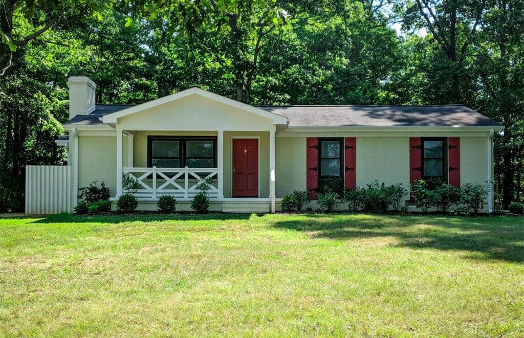 A small, single-story beige house with a red door and shutters. It has a front porch and is surrounded by grass and trees.