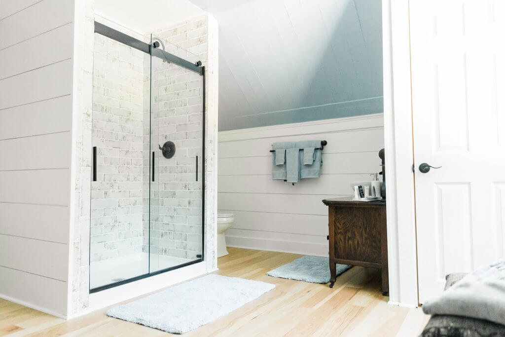 experience oakhaven venues in greensboro 6 Bright bathroom with a glass shower, white brick walls, light wood floors, and blue accents. A wooden cabinet holds toiletries, and towels hang on a black rod.