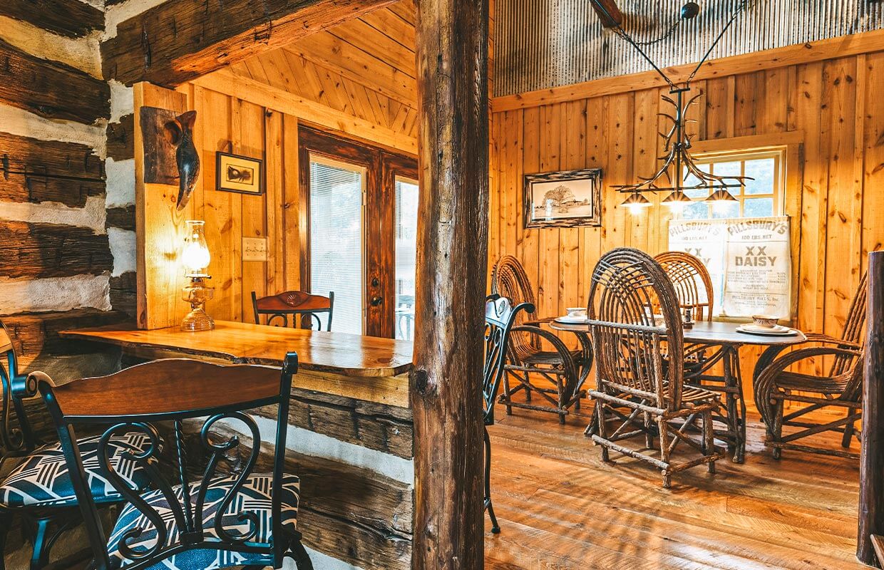 Rustic dining area with wooden walls and floor, wicker chairs around a long table, and decorative items like a mounted fish and framed pictures on the walls.