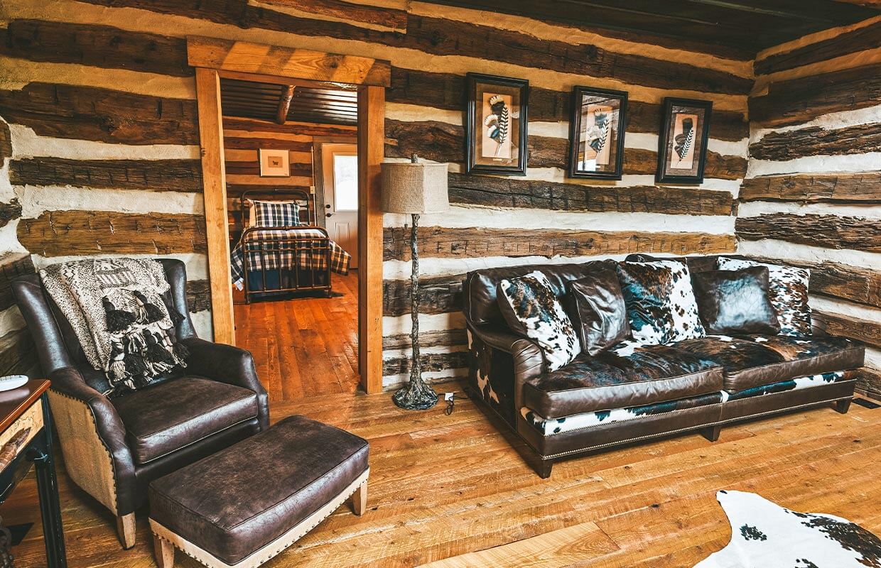 Rustic living room with wooden walls, a leather sofa, and a patterned armchair. A door leading to a bedroom with a checkered bedspread is visible in the background.