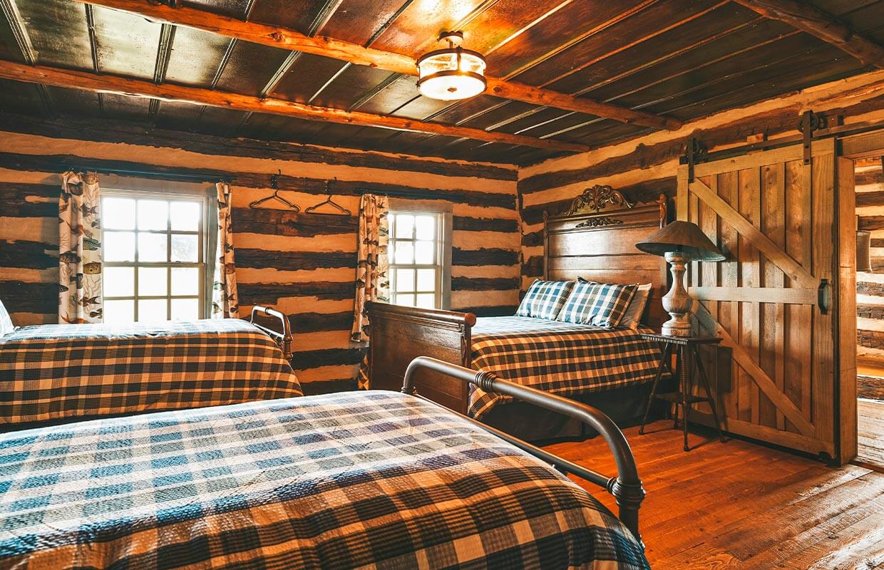 A rustic bedroom with two plaid-patterned beds, wooden walls, a sliding barn door, and two windows with patterned curtains. A lamp sits on a wooden nightstand between the beds.