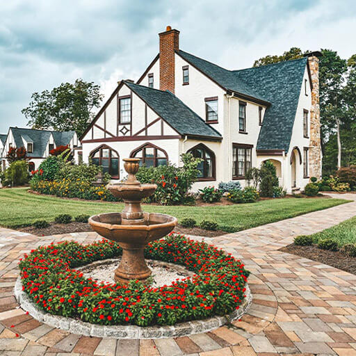 Experience Oakhaven greensboro event venues 18 A two-story Tudor-style house with a dark roof, surrounded by a garden. A circular brick path features a tiered fountain and red flowers in the foreground.