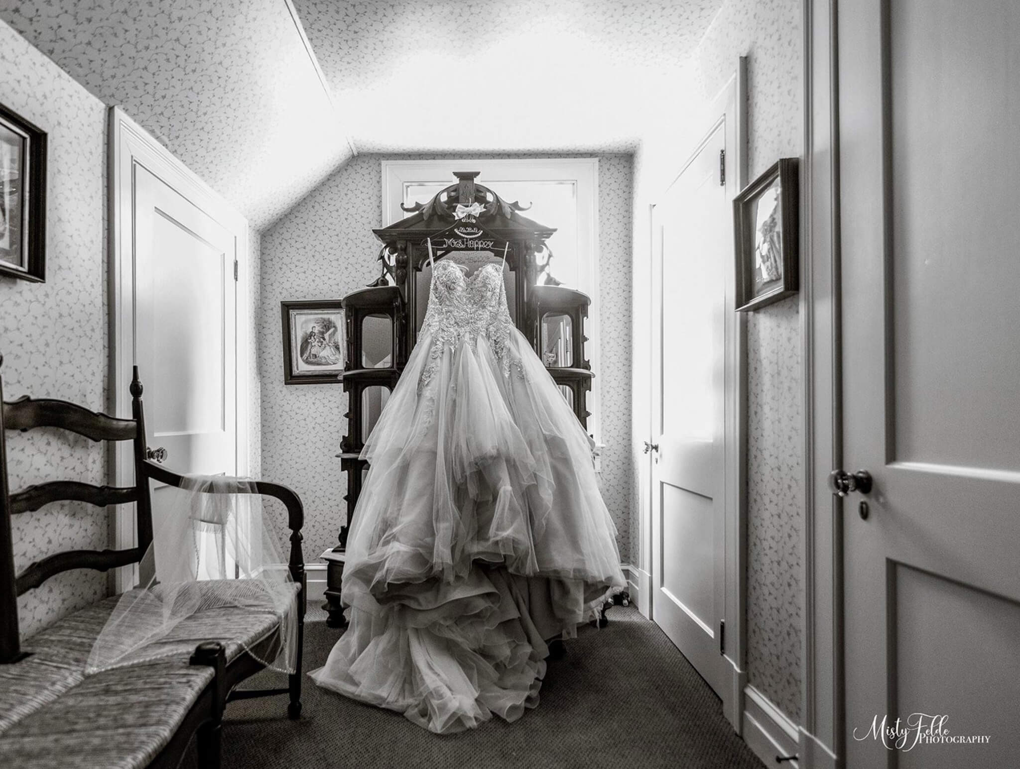 A wedding dress is displayed on a wardrobe in a small, well-lit room with floral wallpaper, a wooden bench, and framed pictures on the walls.