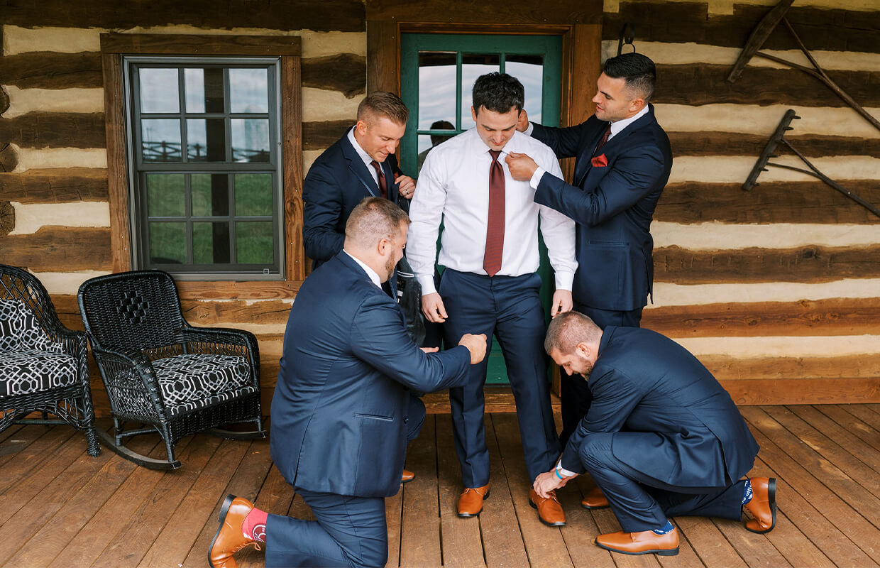 experience oakhaven goldsboro nc wedding venues 21 Five men in suits, three kneeling, adjust and prepare a groom standing on a wooden porch in front of a log cabin.