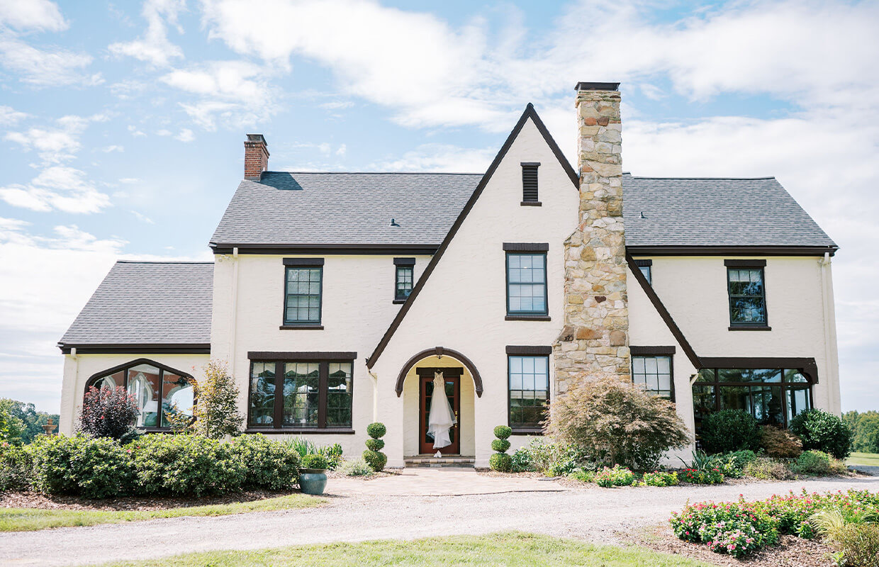 A two-story house with a beige exterior, dark trim, and a large stone chimney is surrounded by greenery under a partly cloudy sky.
