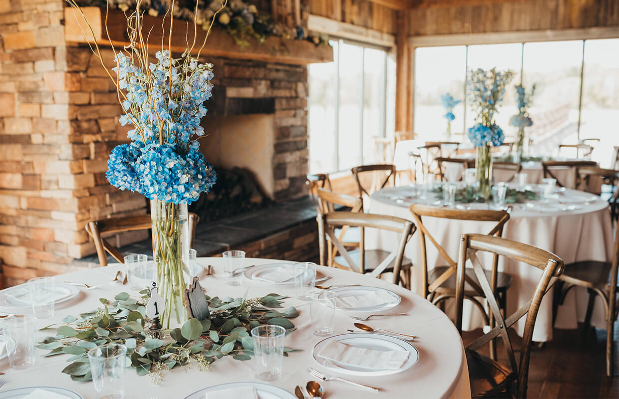 Table setting in a rustic room with round tables covered in white cloths, adorned with glass vases of blue flowers and greenery, surrounded by wooden chairs.