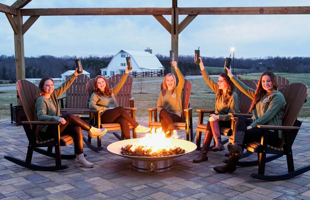 experience oakhaven goldsboro nc wedding venues 5 Five people sitting in Adirondack chairs around a fire pit, raising drinks, with a barn and field in the background at dusk.