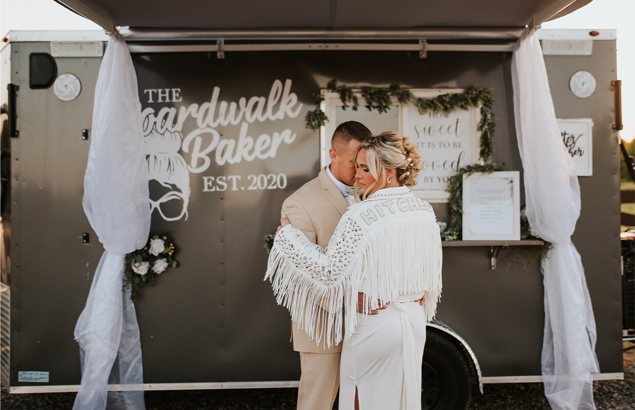 A couple embraces in front of a food truck with "The Boardwalk Baker" sign. The person facing away wears a white fringed jacket with "HITCHED" on the back.