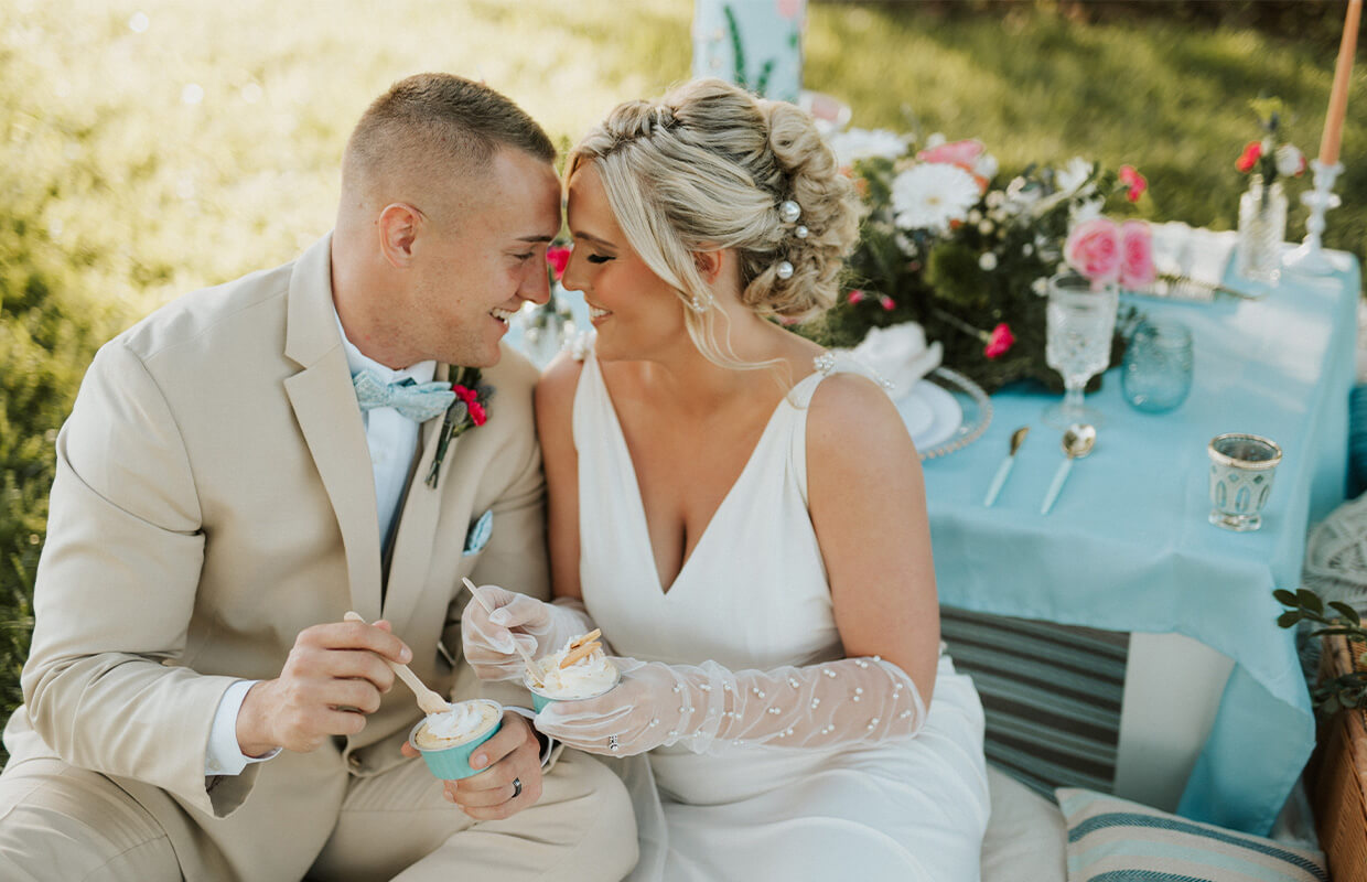 A couple in wedding attire enjoys ice cream while sitting close at a picnic, with a decorated table and flowers in the background.