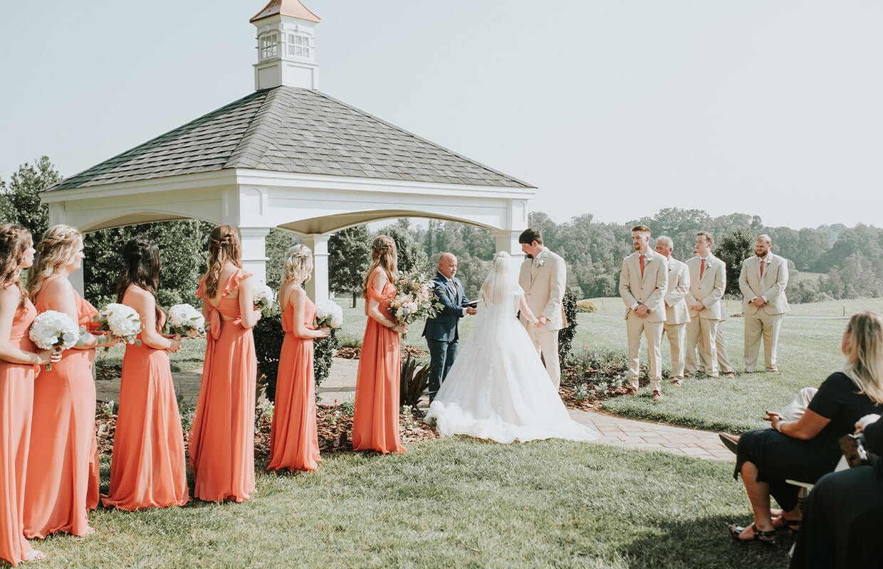 experience oakhaven greensboro nc wedding venues 18 A wedding ceremony outdoors with a couple at the altar. Bridesmaids in coral dresses stand to the left; groomsmen in beige suits to the right. A gazebo and green landscape are in the background.
