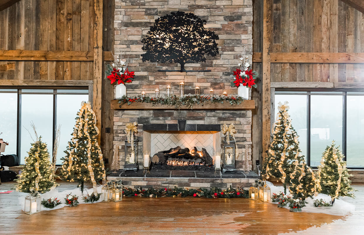 experience oakhaven greensboro nc wedding venues 17 A rustic stone fireplace decorated with candles, garlands, and red flowers. Christmas trees with lights and ribbons surround the fireplace. Large windows on both sides.