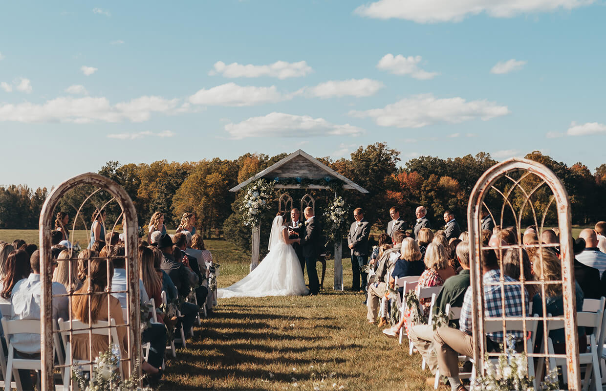 Blog8experience oakhaven greensboro nc wedding venues 14 Outdoor wedding ceremony with a couple exchanging vows under a wooden arch adorned with flowers. Guests are seated on both sides, and the setting features a grassy field with a forested backdrop.