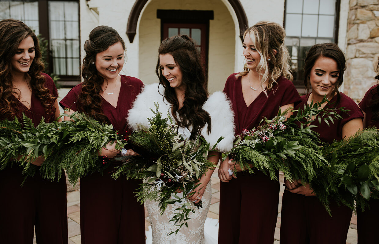 Bride in a white fur shawl stands with four bridesmaids wearing maroon dresses, all holding green bouquets, in front of a building.