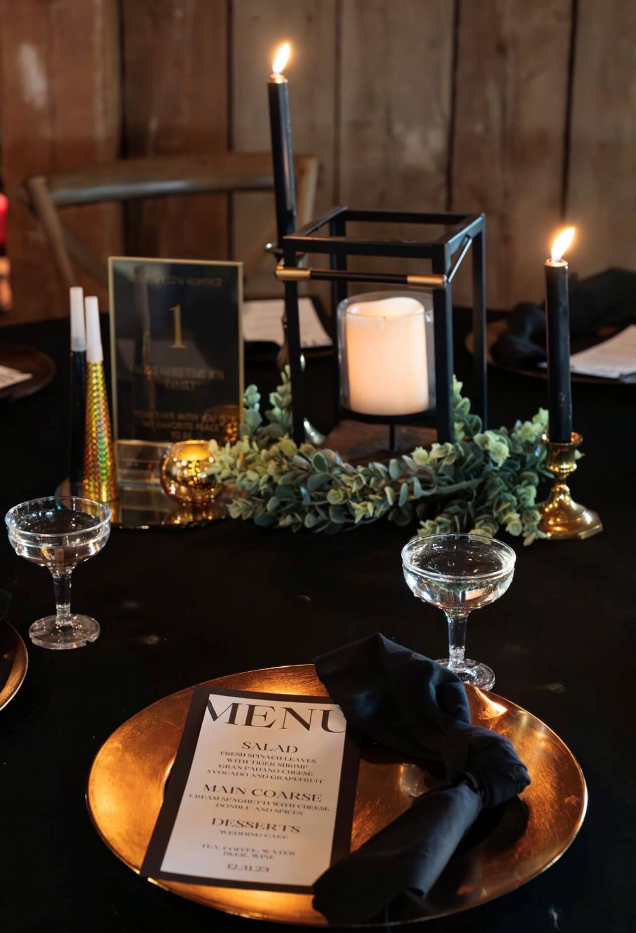 Elegant table setting with a gold plate, menu, black napkin, and candle centerpiece on a dark tablecloth. Two black candles and two champagne glasses are also present.