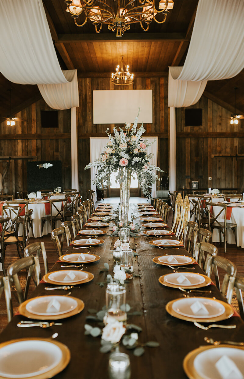 Rustic banquet hall with a long wooden table set for a meal, featuring elegant floral centerpieces, gold-rimmed plates, and white napkins under chandeliers and draped fabric.