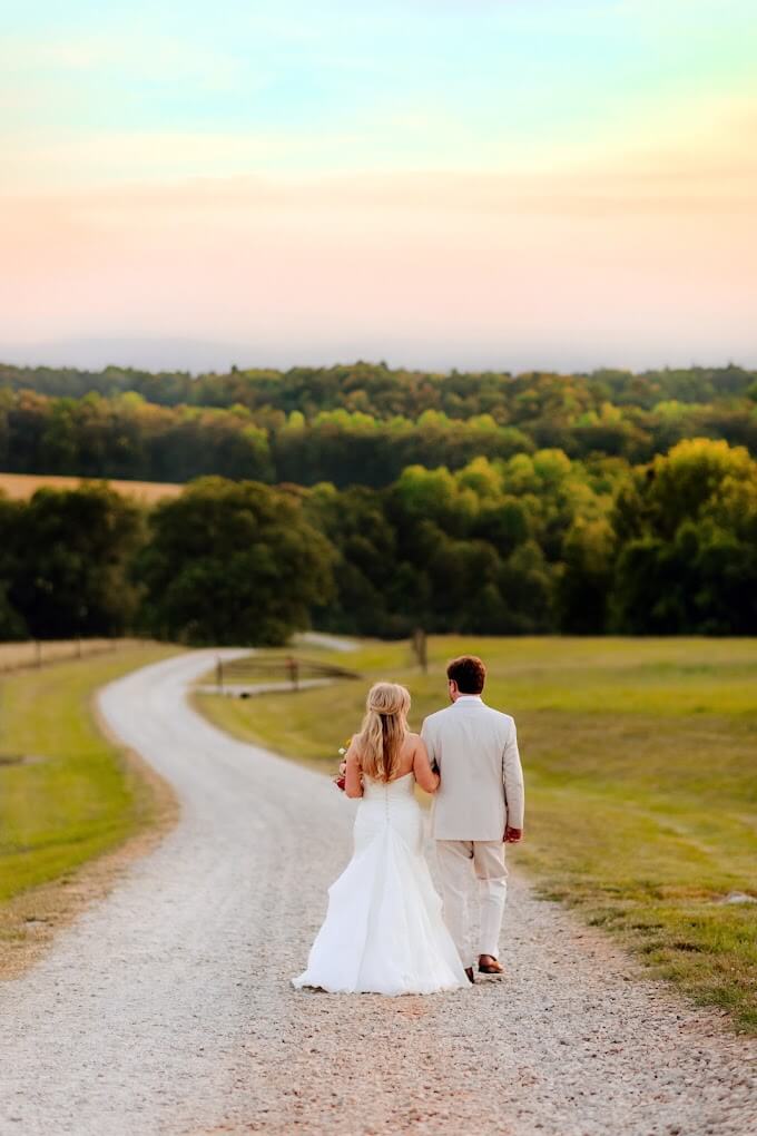 Experience Oakhaven wedding venues in nc 10 A bride and groom walk hand in hand down a country road at sunset, surrounded by green fields and trees.