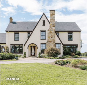 A large two-story manor house with a beige exterior, dark trim, and a prominent stone chimney, surrounded by a manicured lawn and shrubs.
