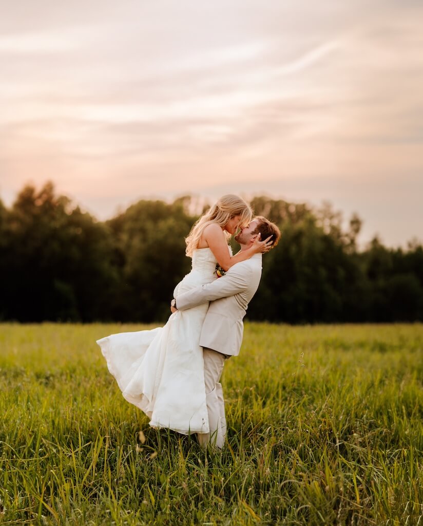 A groom in a light suit lifts a bride in a white dress in a grassy field at sunset. Trees are in the background.