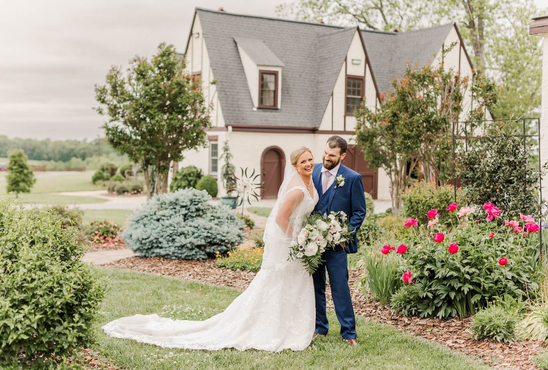 Bride and groom standing together in a garden, with a house in the background and colorful flowers surrounding them.