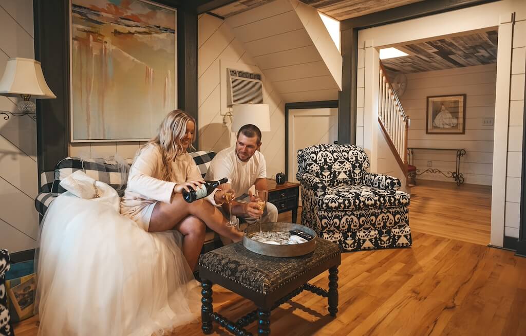 Experience Oakhaven North carolina Wedding Venue 21 A couple sits on a couch in a cozy, wood-floored living room. They are pouring wine into glasses. The room features a patterned armchair, a side table, and a large painting on the wall.