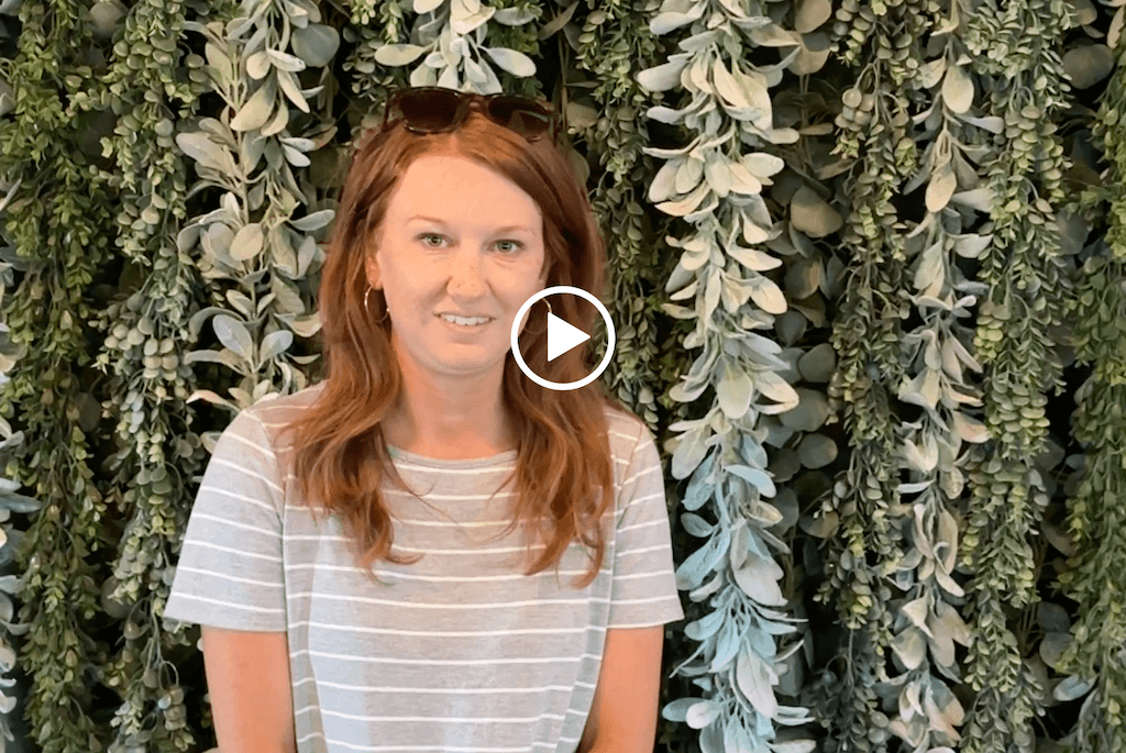 A person with long hair and a striped shirt stands in front of a leafy green wall with sunglasses on their head.
