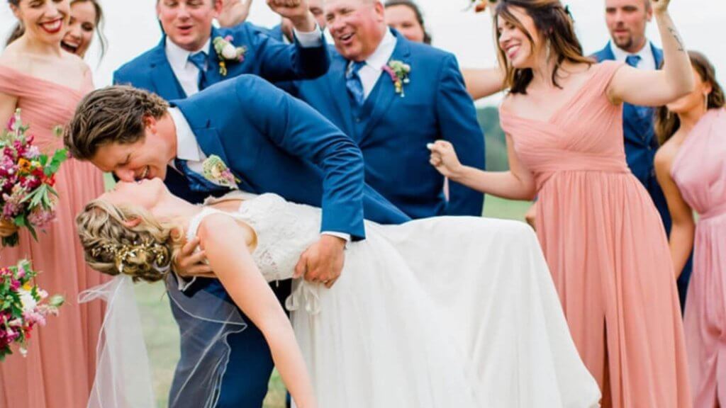 A groom in a blue suit dips and kisses the bride in a white dress. Bridesmaids in pink dresses and groomsmen in blue suits cheer behind them.