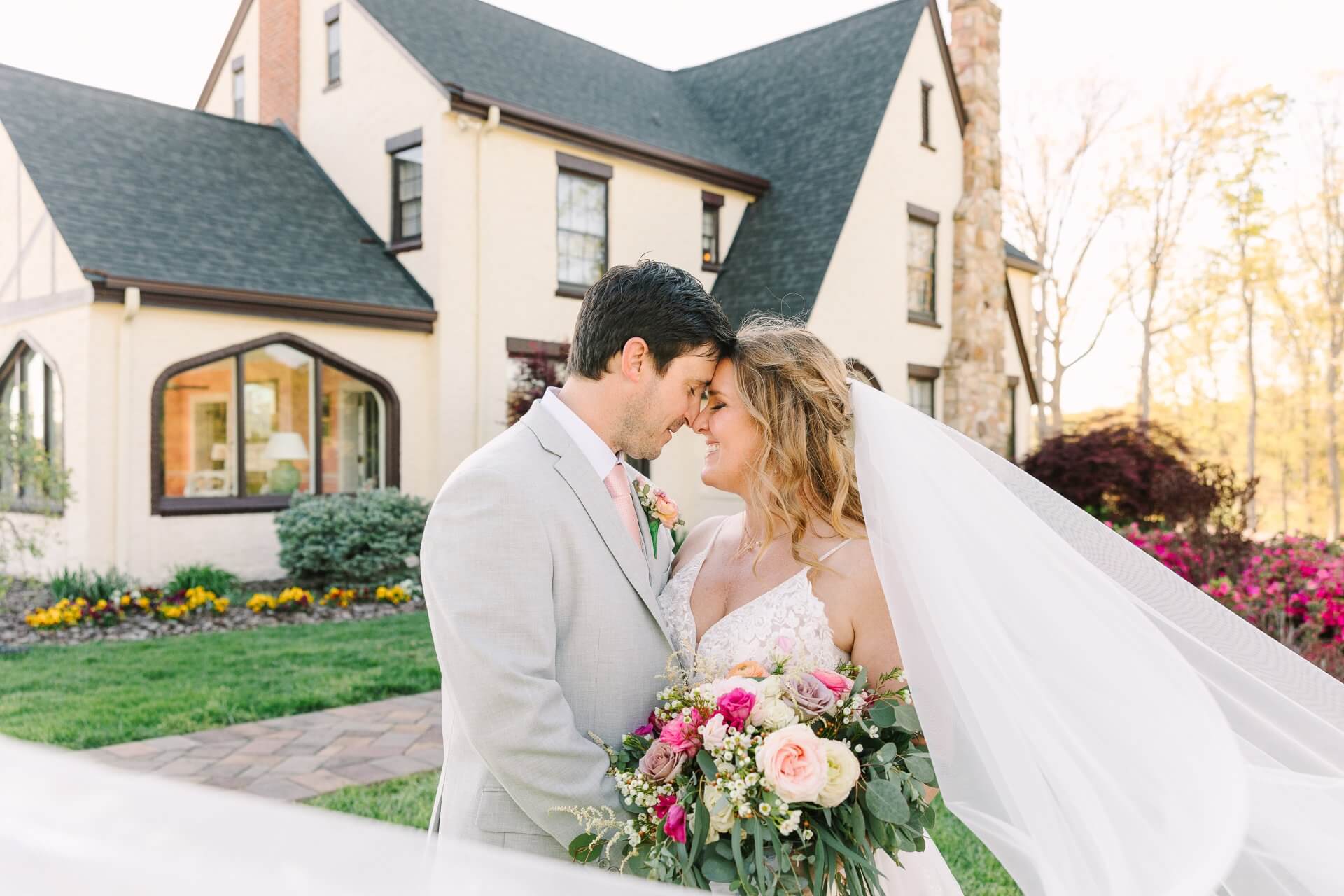 Experience Oakhaven NC Wedding Venue 19 A bride and groom embrace in front of a house, surrounded by flowers. The bride holds a bouquet and her veil flows in the breeze.