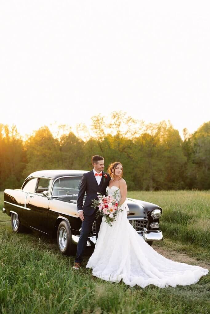 Bride and groom stand beside a classic black car in a field at sunset. The bride holds a bouquet, wearing a white gown, and the groom is in a dark suit with a red bow tie.