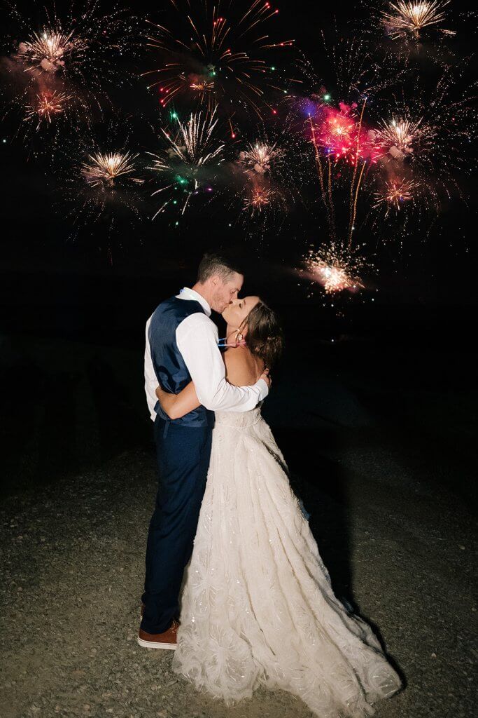 A couple in wedding attire kisses under a night sky filled with colorful fireworks.