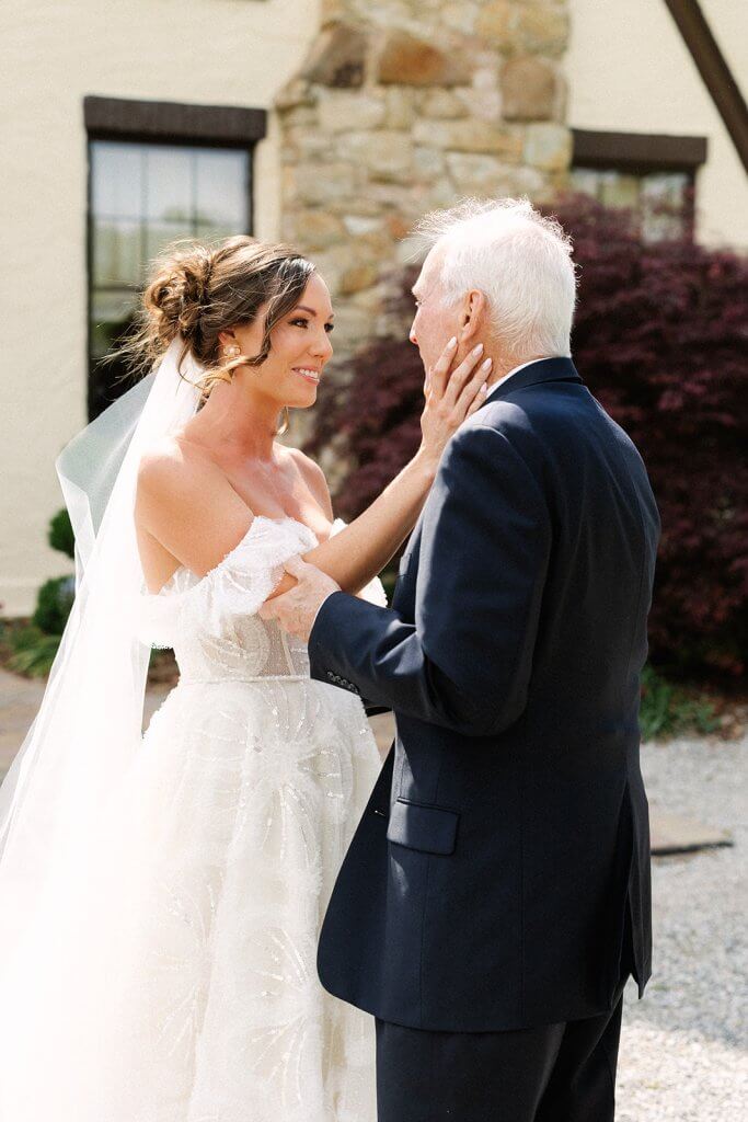 A bride in a white dress and veil smiles and holds the face of an older man in a suit outside a building.