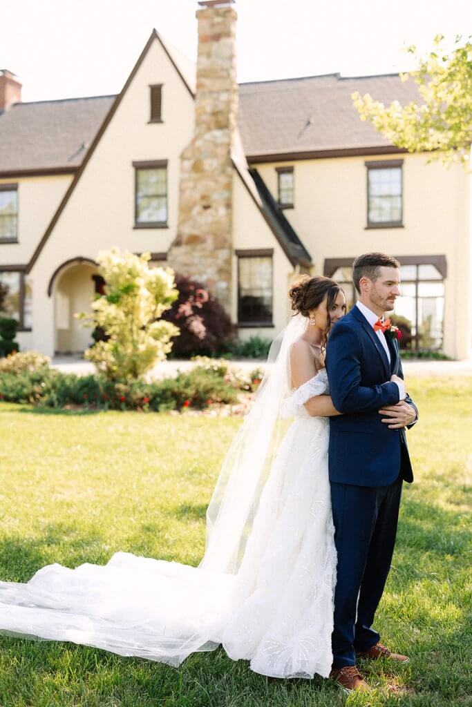 A bride in a white dress and veil stands with a groom in a blue suit outside a large house.