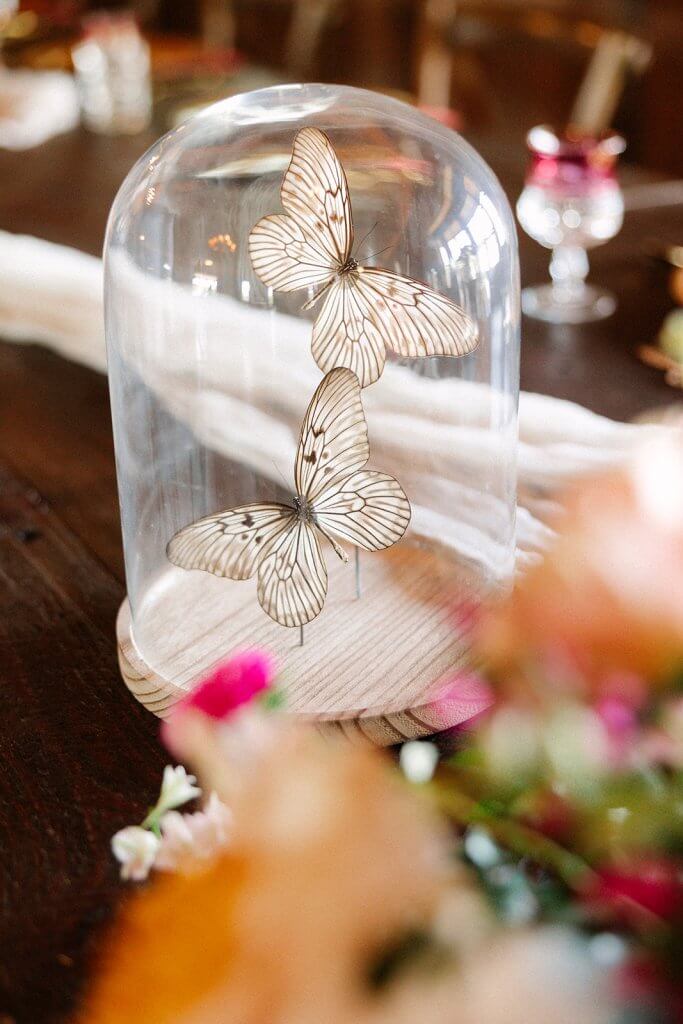 Two decorative glass butterflies are displayed under a clear dome on a wooden base, surrounded by soft-focus pink and orange flowers.