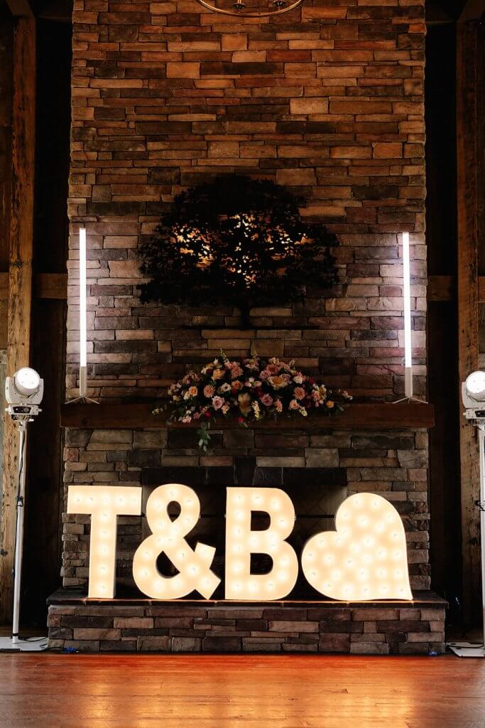 Large lighted letters "T&B" with a heart symbol adorn a stone fireplace, surrounded by floral arrangements and vertical lights, creating the perfect ambiance for a spring wedding.