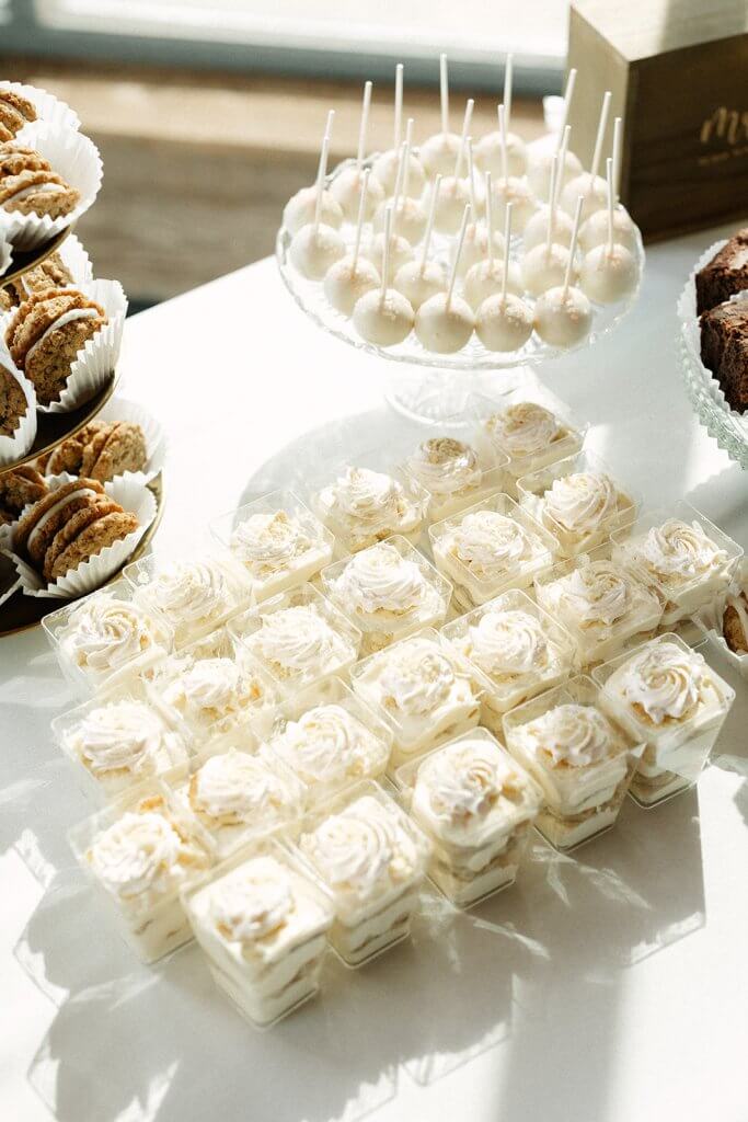 An assortment of desserts including cake pops, mini cupcakes in plastic cups with cream topping, and cookies displayed on a white table.