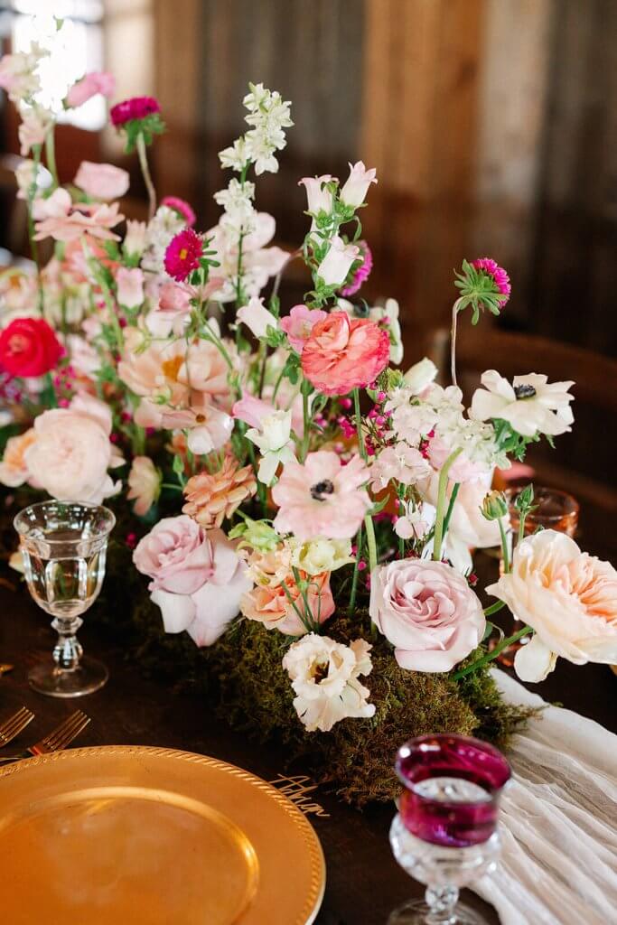 A table centerpiece with pink and white flowers, greenery, and moss, surrounded by glassware and a gold plate on a wooden table.