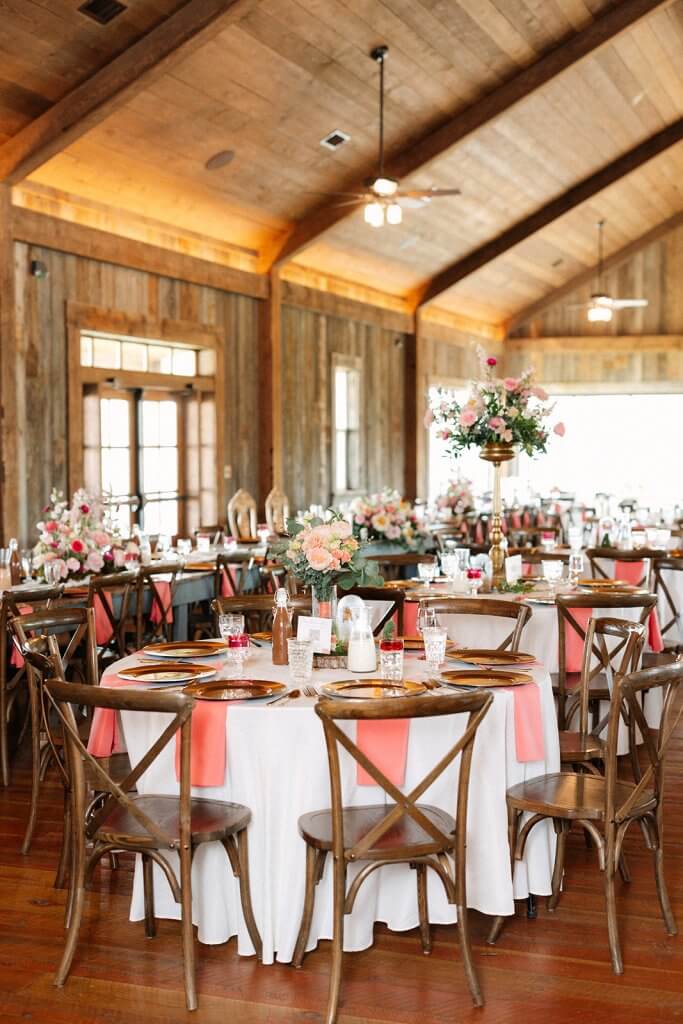 A rustic banquet hall with wooden chairs and tables set in white and pink decor. Floral centerpieces adorn the tables, and natural light filters through large windows.
