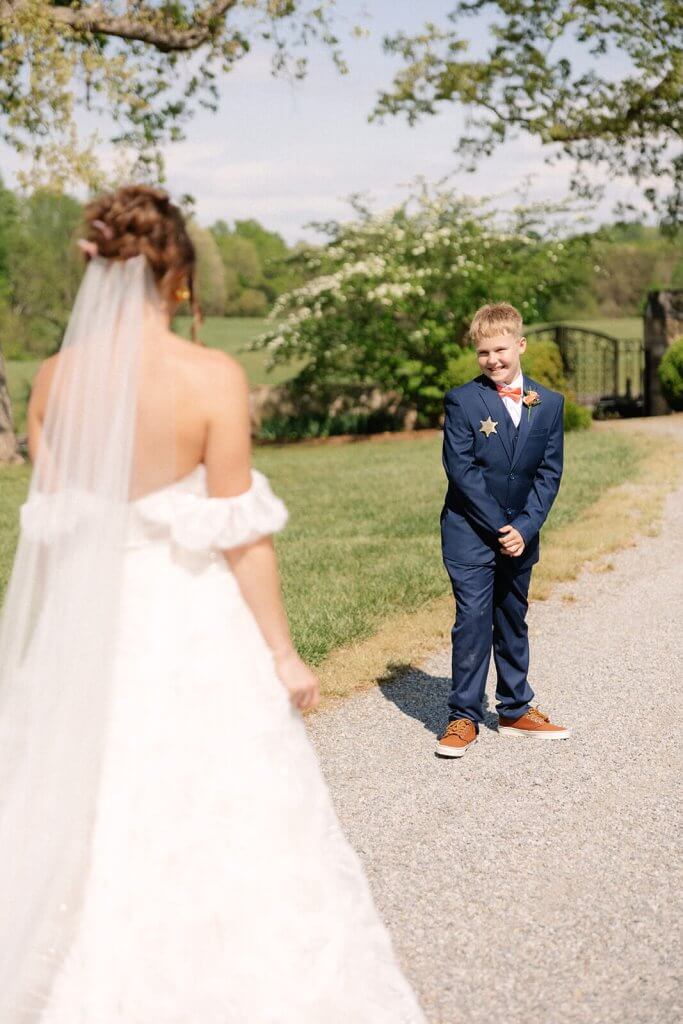 A bride and a young boy in a suit stand facing each other on a gravel path. The boy is smiling, and the background features grass, trees, and a white flower bush.