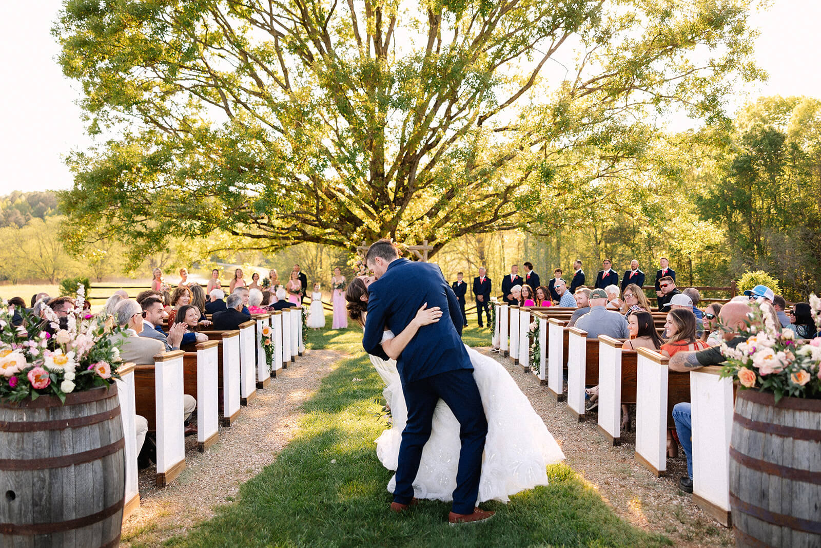At a charming spring wedding, a couple shares a kiss under a large tree during their outdoor ceremony, surrounded by guests seated on wooden benches adorned with delicate flower arrangements.