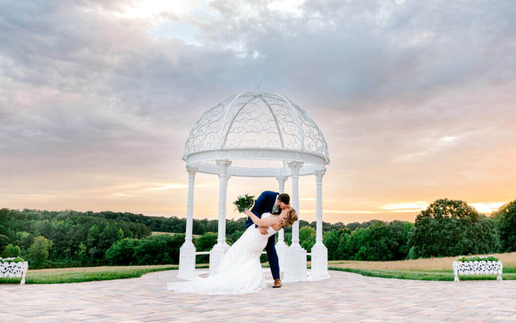 A couple in wedding attire kisses under a white gazebo at sunset during their outdoor wedding ceremony, with green trees and a cloudy sky in the background.