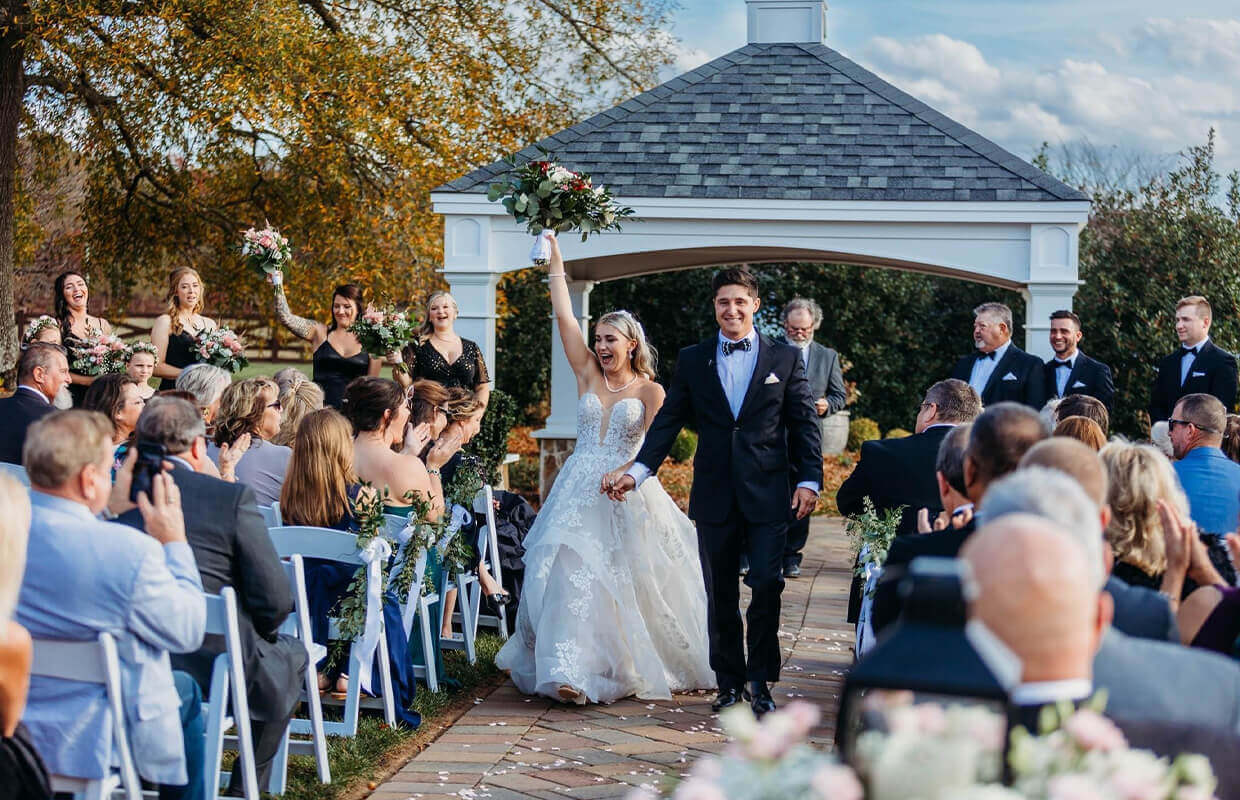 A bride and groom walk down the aisle, holding hands and smiling, as wedding guests seated on either side applaud under a gazebo during a sunny outdoor wedding ceremony.