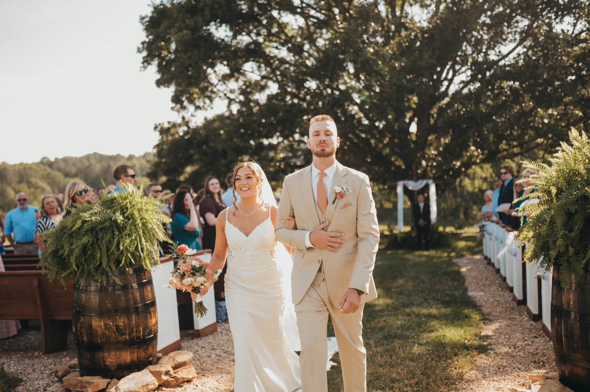 A bride and groom walking down an outdoor ceremony aisle with ferns.