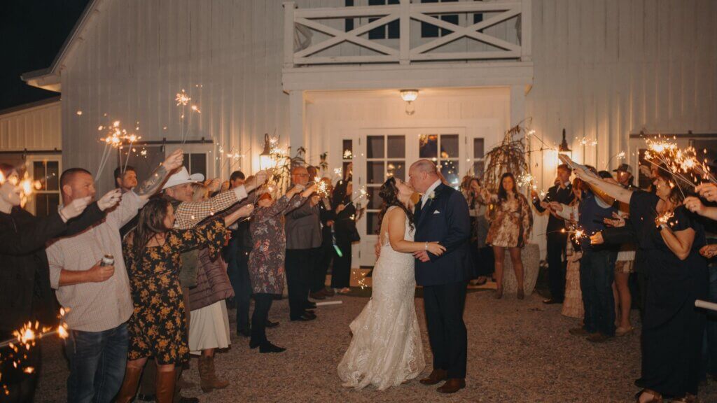 A bride and groom kiss outside a barn venue at night, surrounded by guests holding sparklers in celebration.