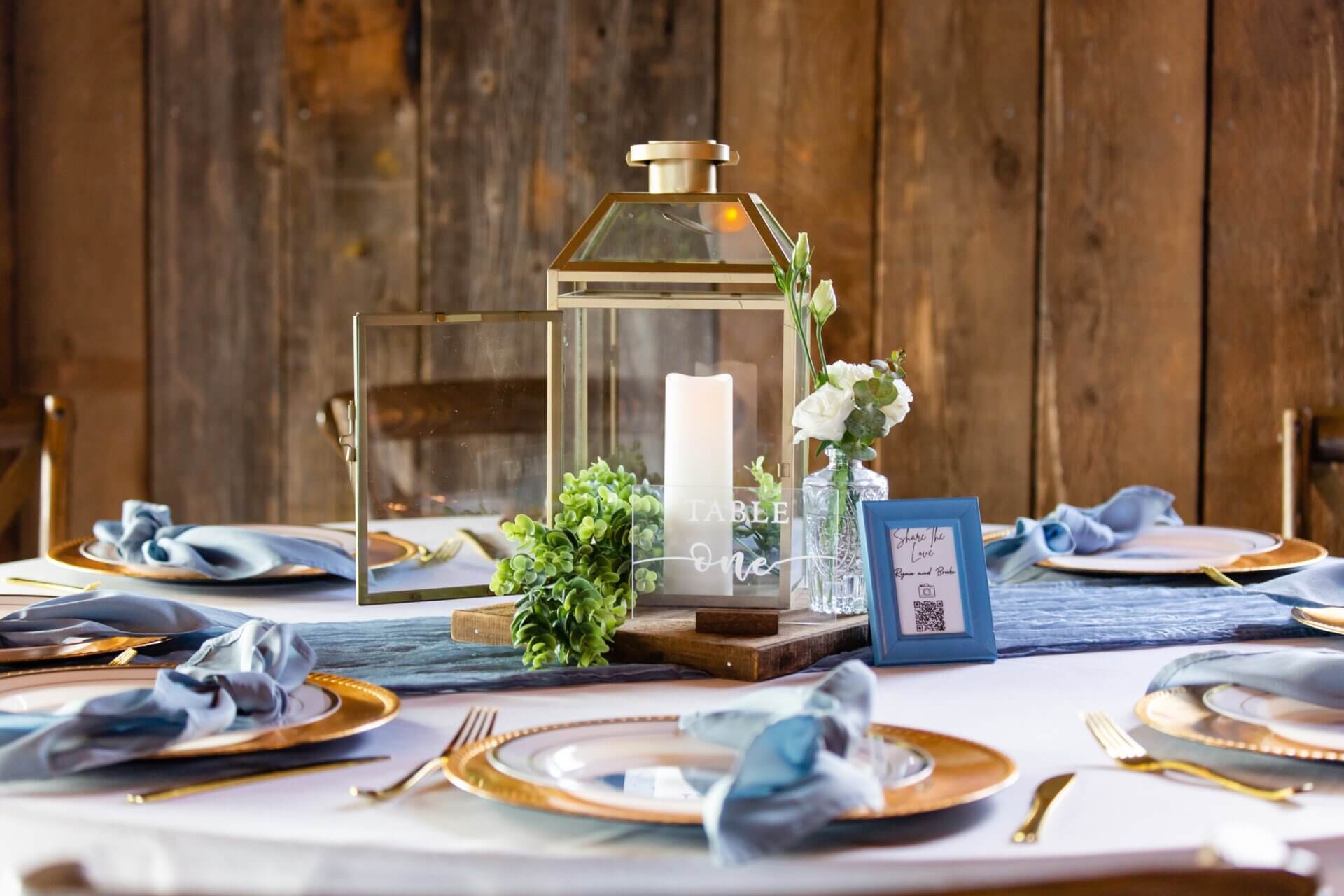 A table setting with a lantern and flameless candle adorned with greenry complementing blue custom linens.