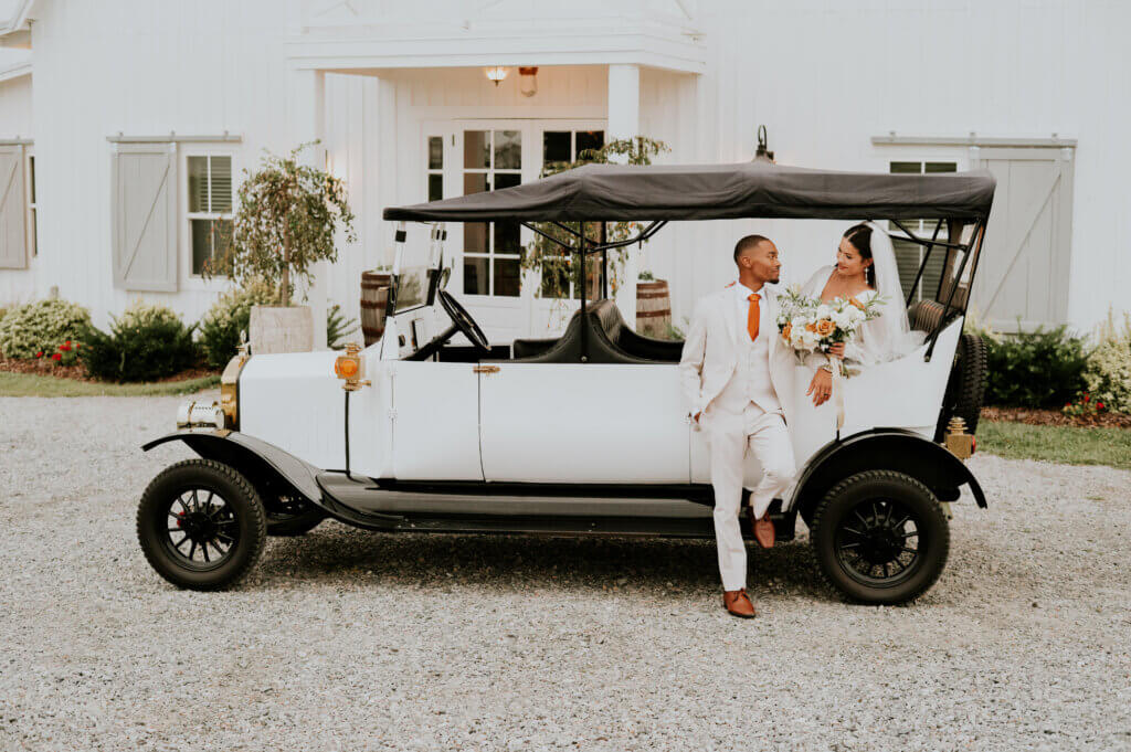 A bride and groom with wedding venue Oakhaven's signature Model T golf cart in front of the entrance to the white barn venue.